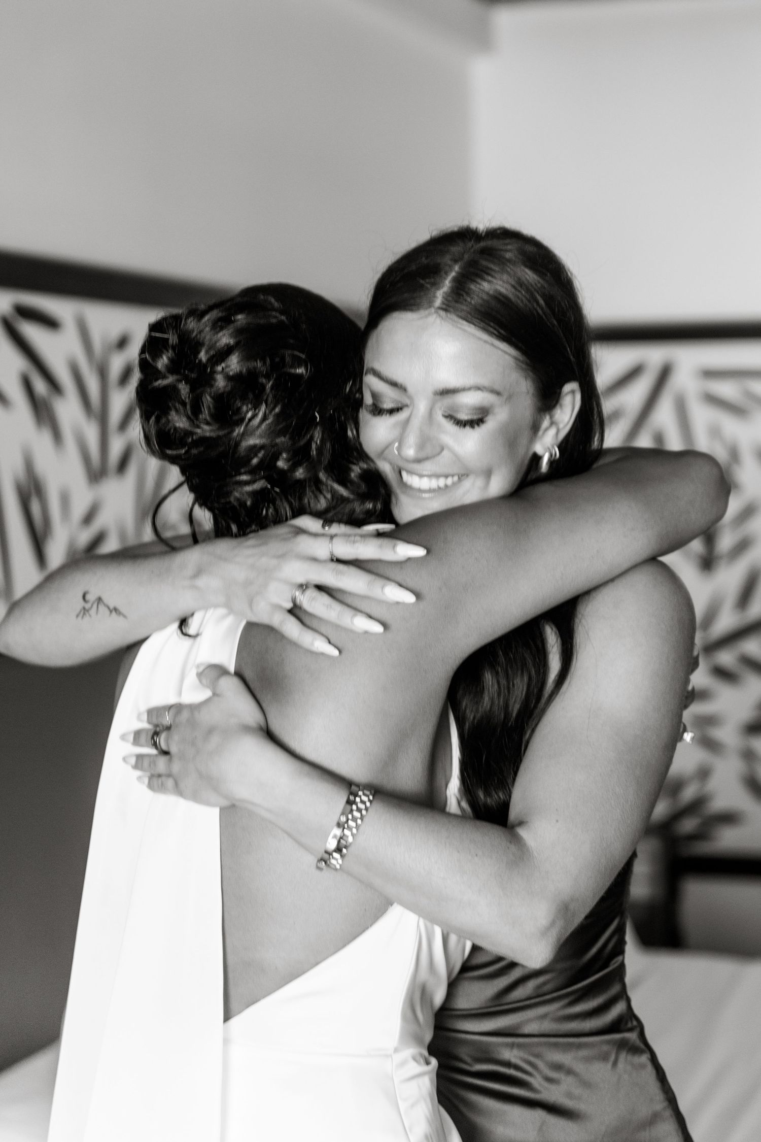 Bride in white gown receives warm hug from smiling bridesmaid in black and white wedding photo.