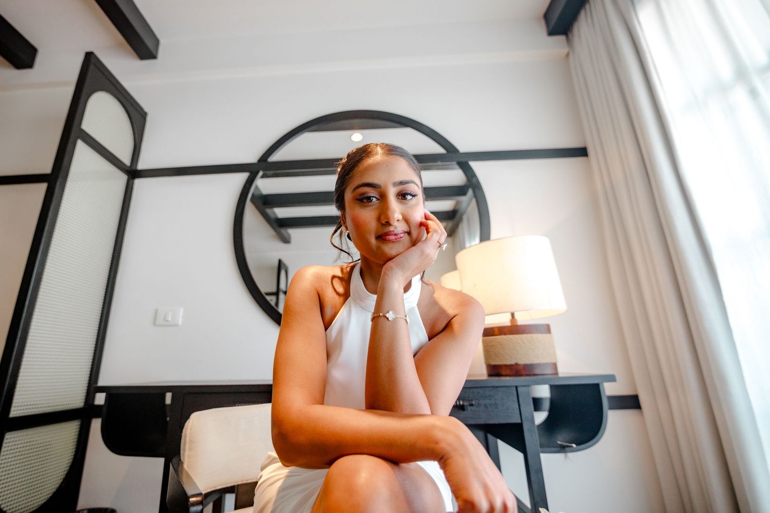 Young woman in white dress sitting thoughtfully in elegant hotel room with round mirror and lamp.