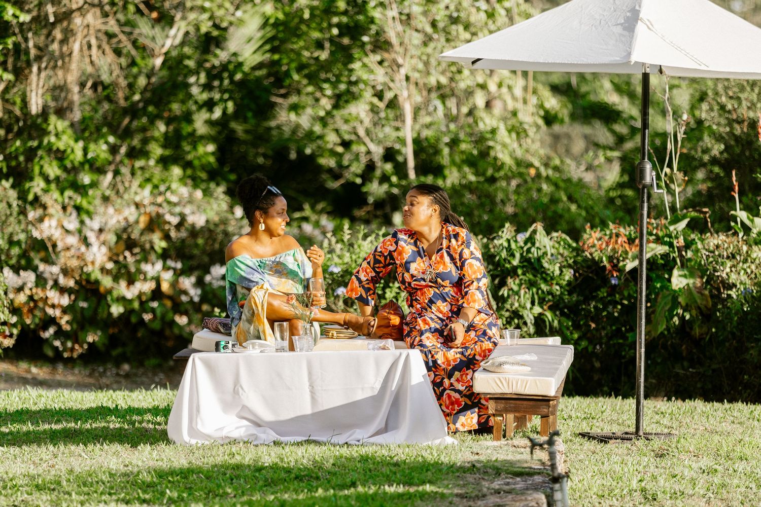 Two people in colorful floral outfits sitting at white-draped table under umbrella in garden.