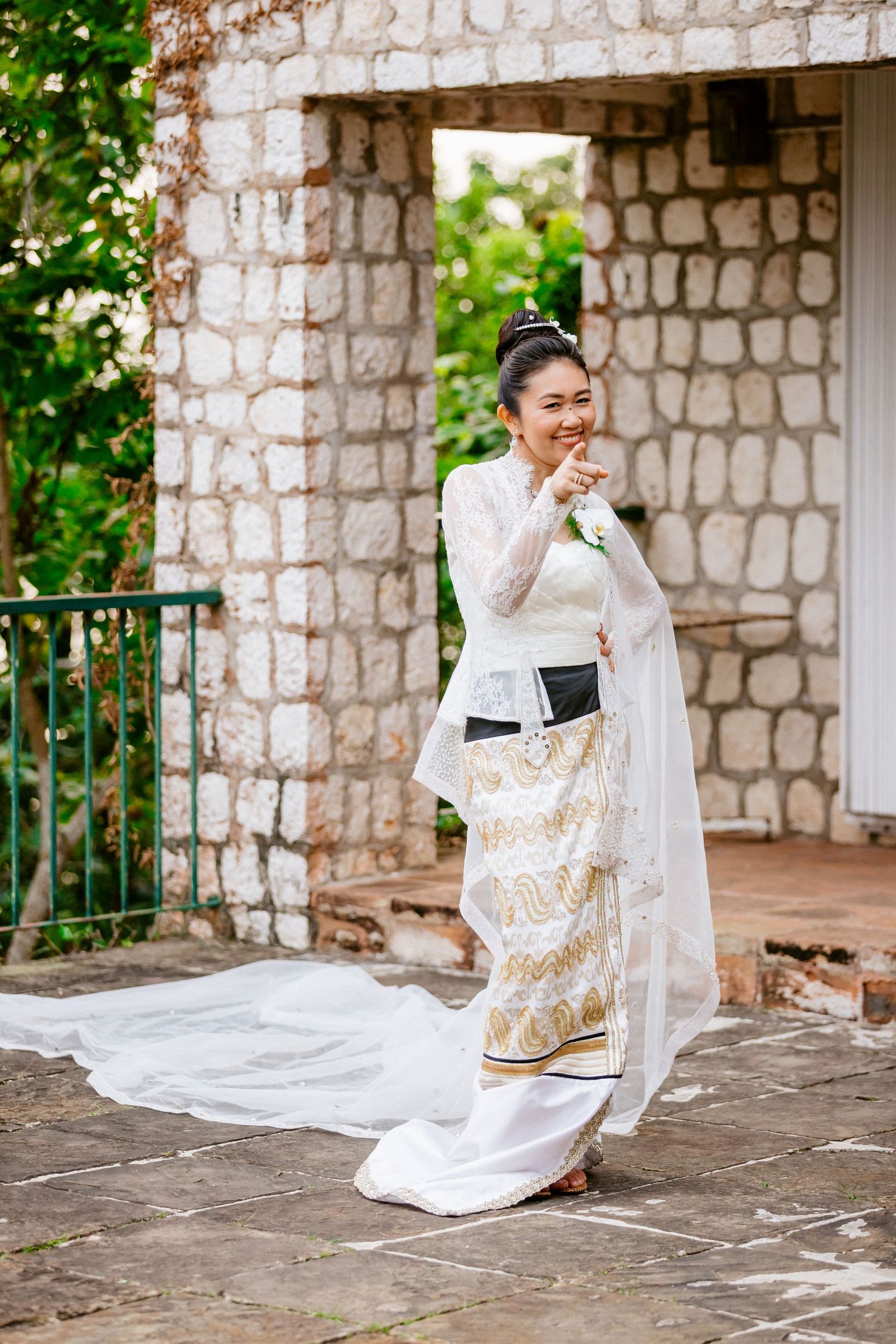 Smiling Asian bride in elegant white and gold traditional wedding dress standing outdoors by stone pillars.