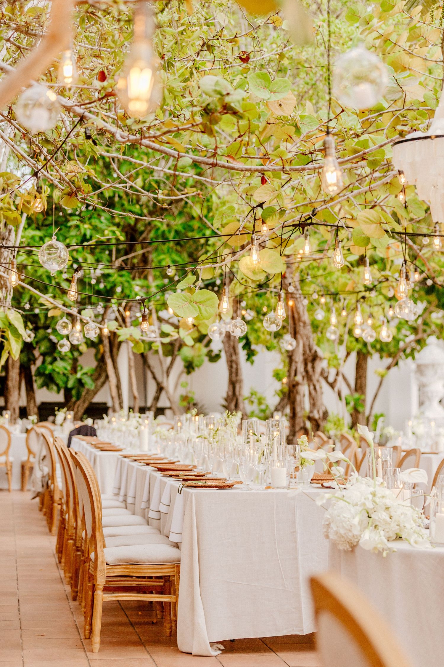 Magical outdoor wedding reception with Edison bulb string lights, long white linen tables, and white floral centerpieces.