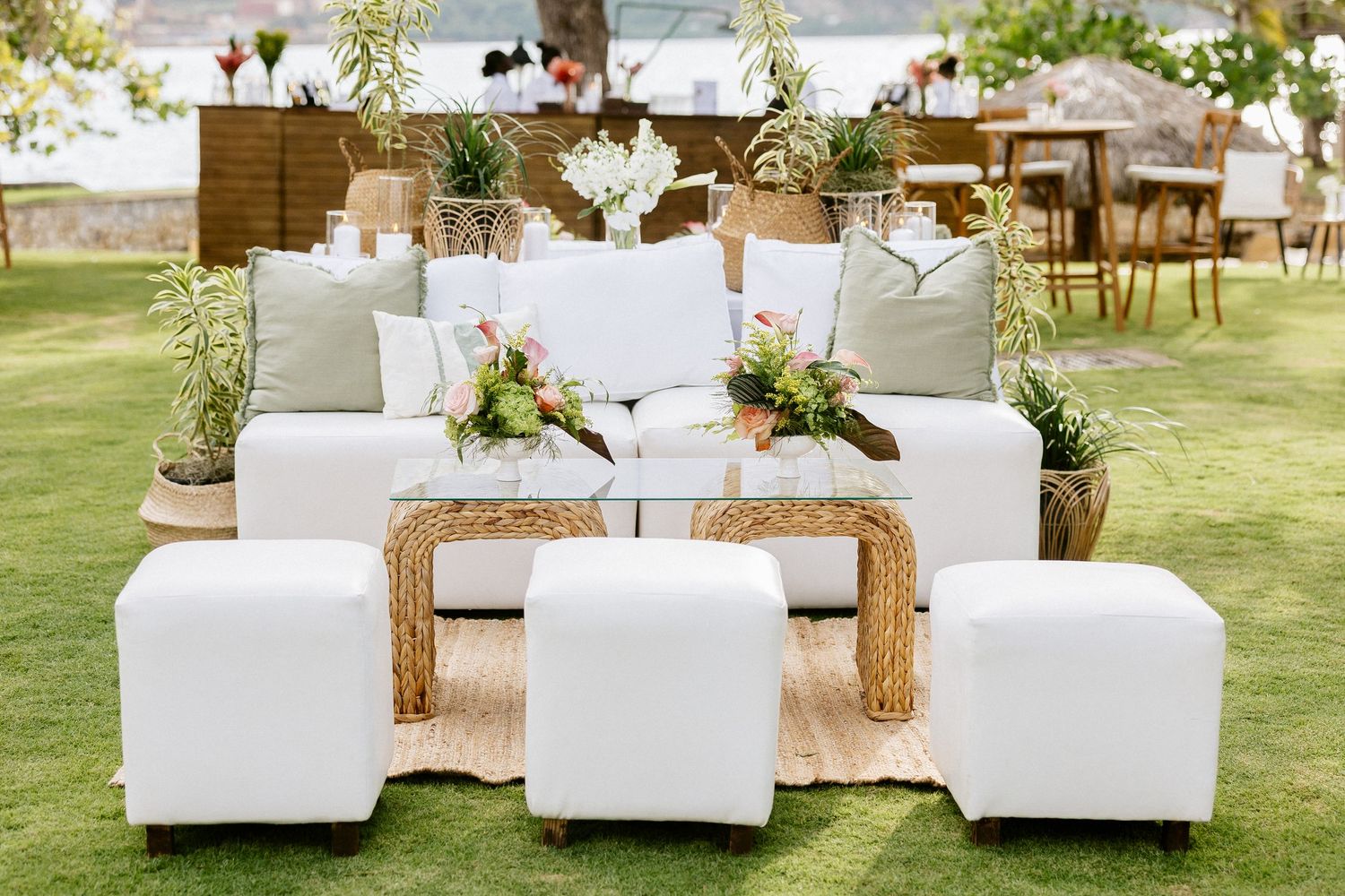 Elegant outdoor wedding lounge with white sofa, ottomans, wicker coffee table, and floral arrangements on green lawn.