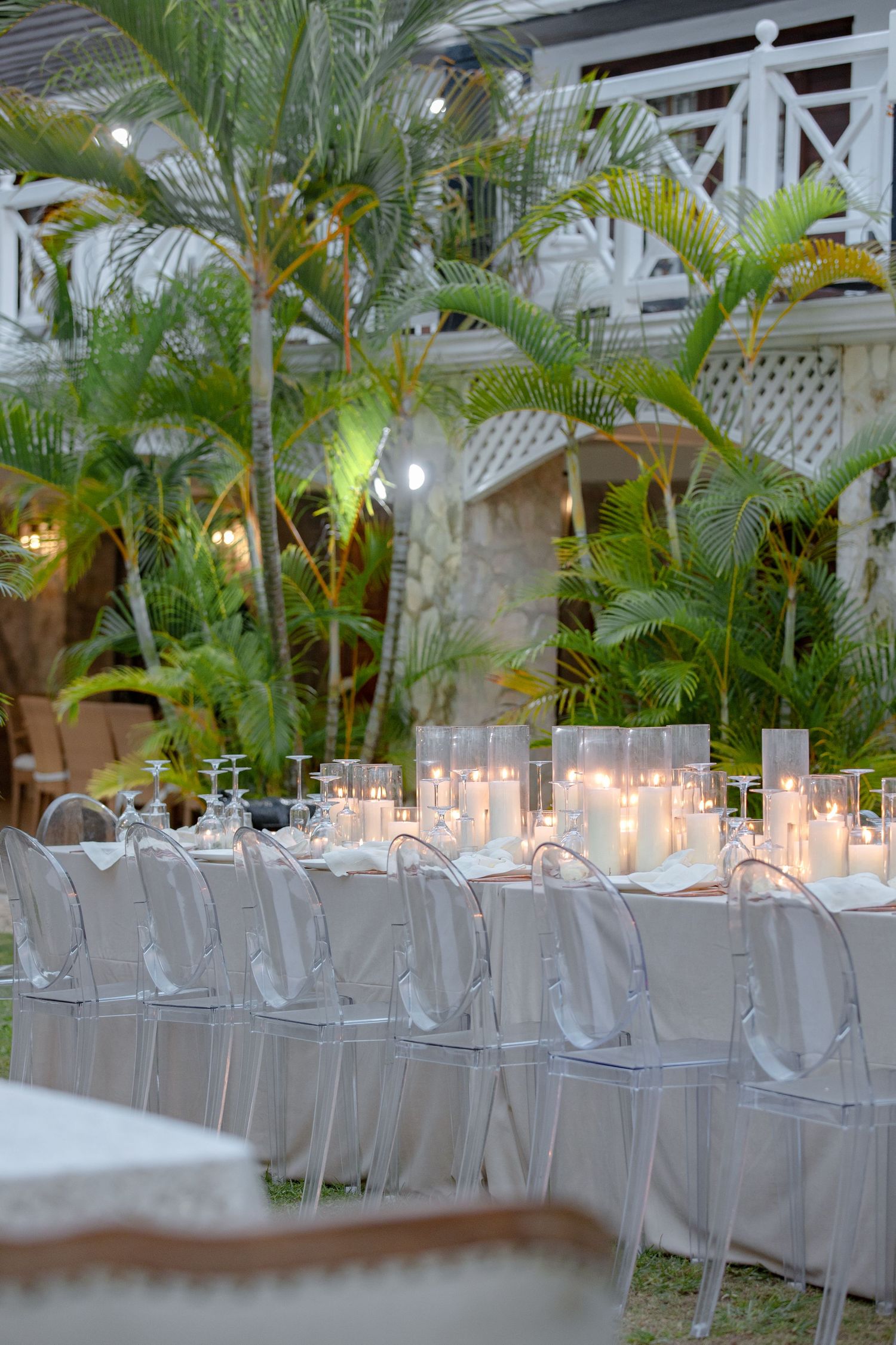 Outdoor evening wedding reception with ghost chairs, candlelit table, and tropical palm tree backdrop.