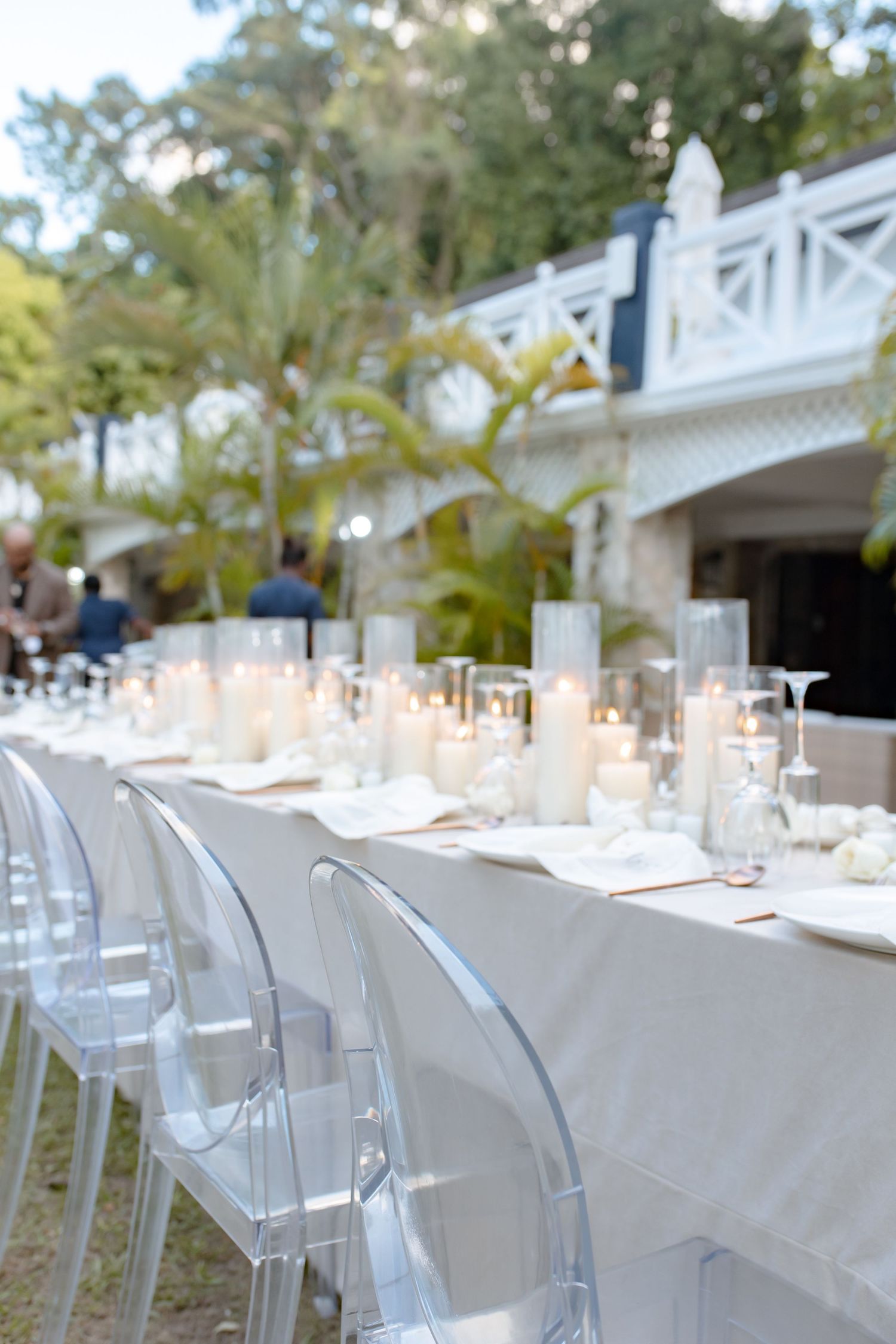Outdoor wedding reception long table with white linens, pillar candles, and ghost chairs surrounded by tropical greenery.