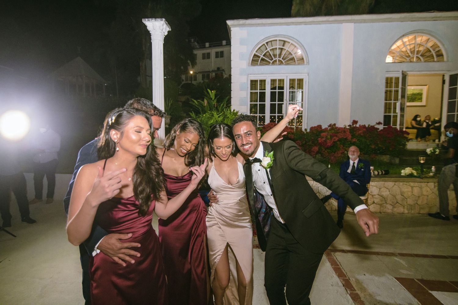 Groom taking a joyful selfie with bridesmaids in burgundy gowns outside an elegant venue at night.