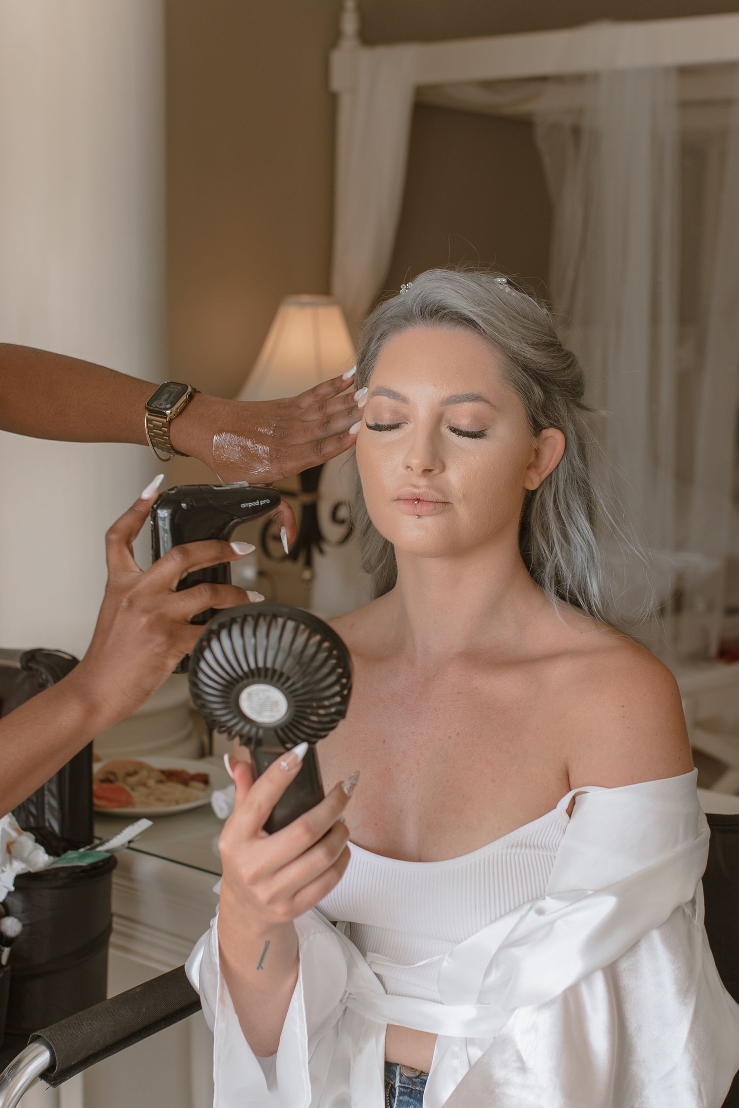 Bride with silver hair getting makeup applied while holding a small fan mirror.