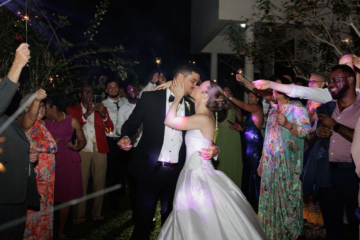 Bride and groom share a romantic kiss surrounded by guests holding sparklers at their nighttime wedding reception.