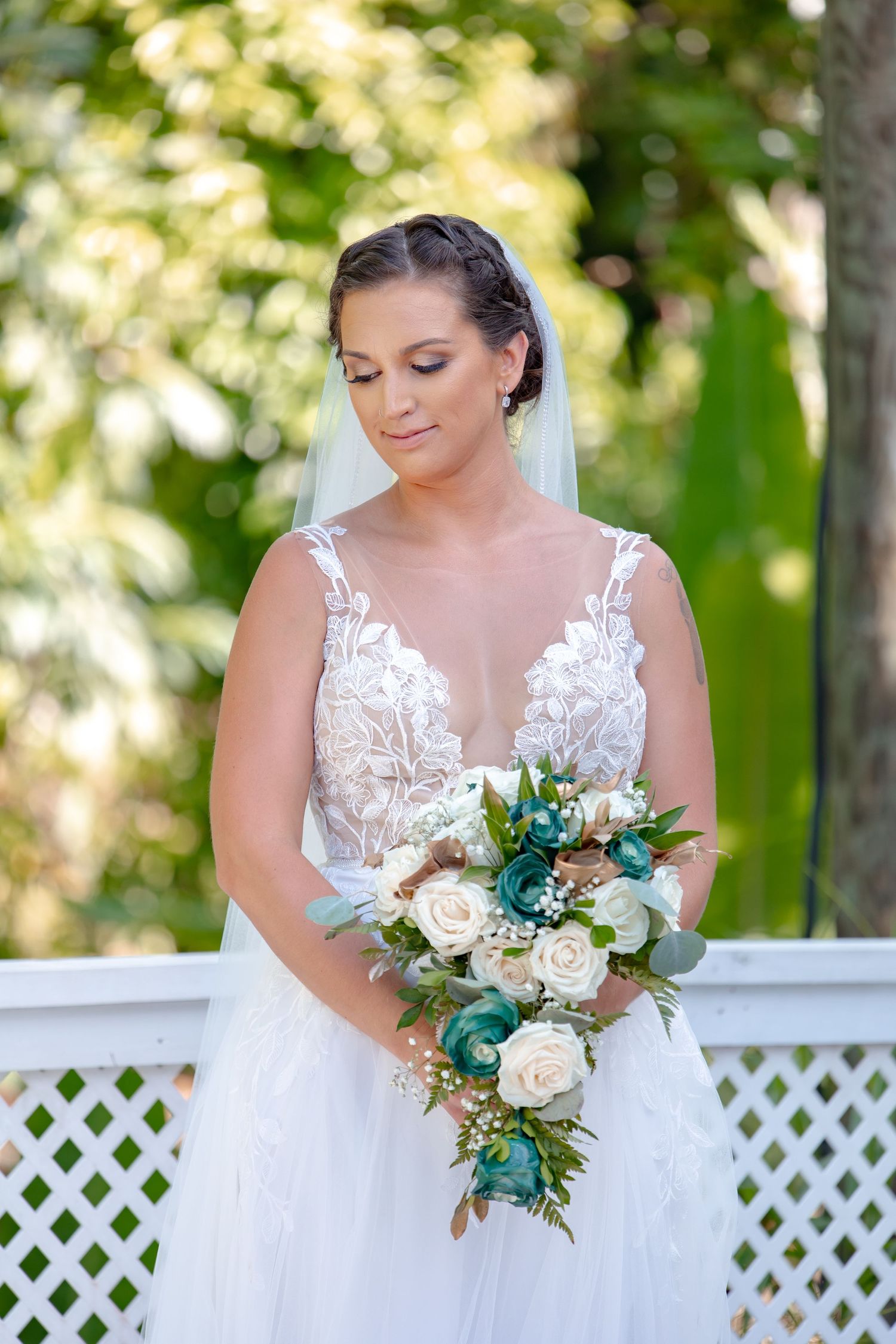 Bride in lace wedding gown holding teal and white bouquet while standing on outdoor white deck.