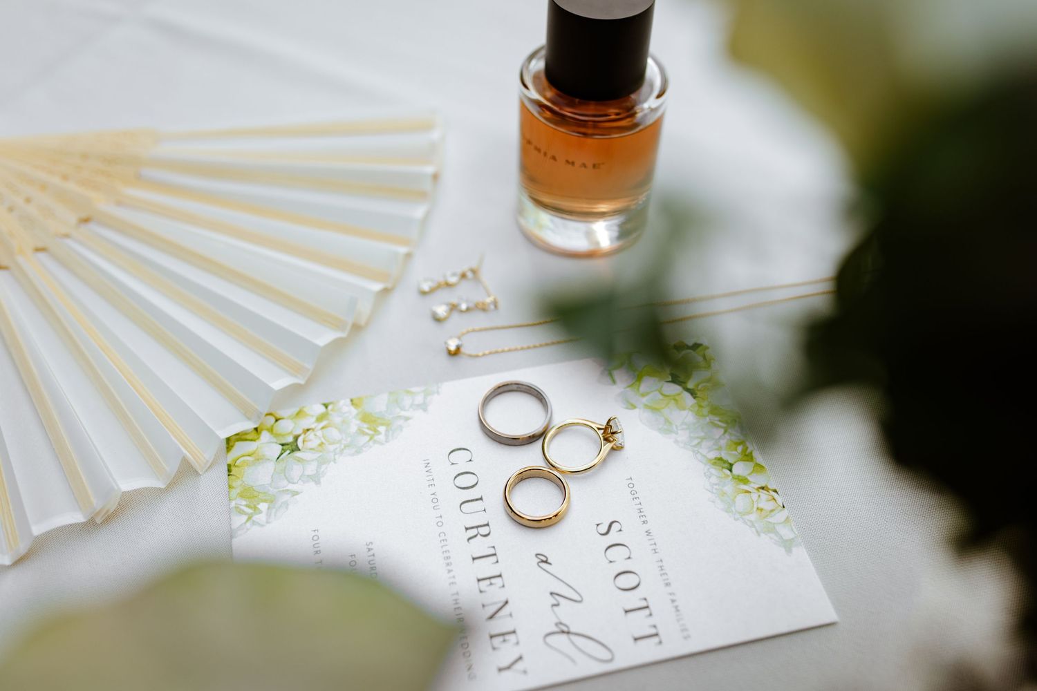 Wedding details flat lay with gold bands, floral stationery, white paper fan, and amber perfume bottle.