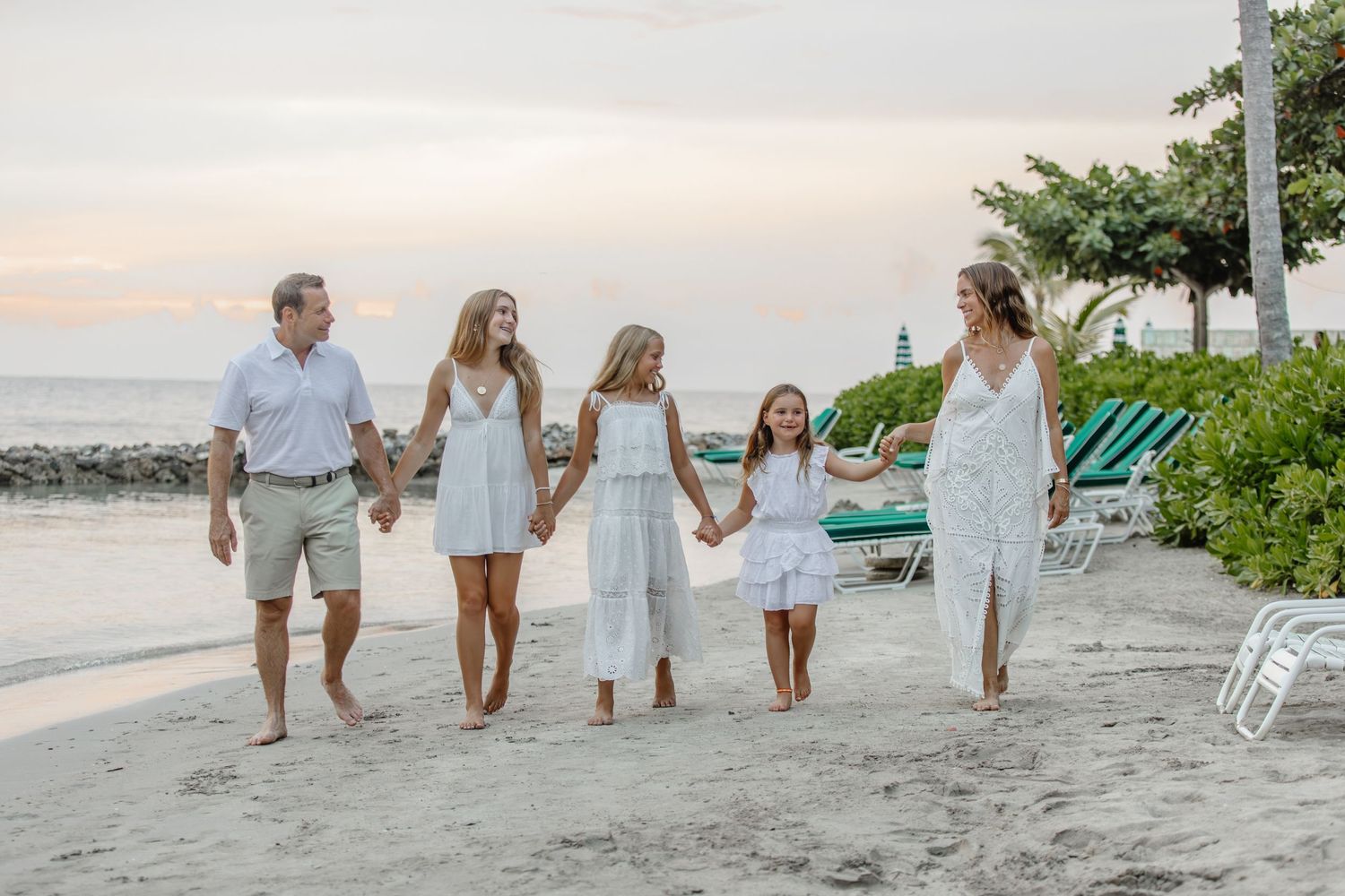 Family of five in white outfits walking hand-in-hand on a sandy beach at sunset near tropical greenery.