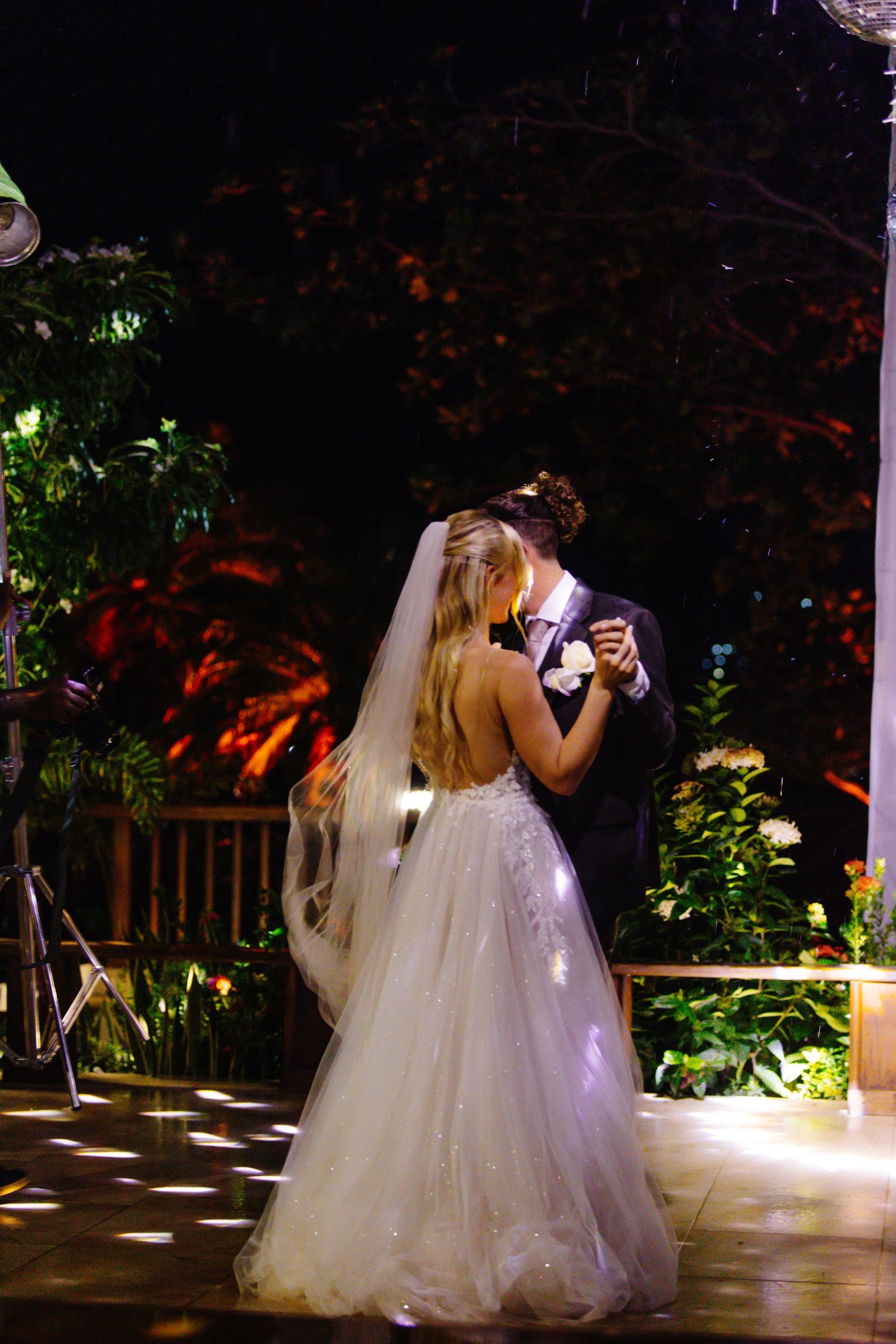 Bride and groom share their first dance outdoors at night, surrounded by lush greenery and warm lighting.