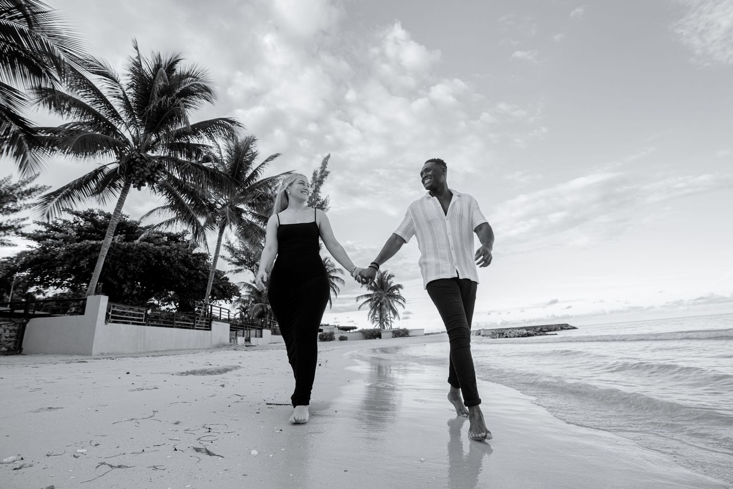 Black and white photo of couple holding hands walking on a tropical beach.