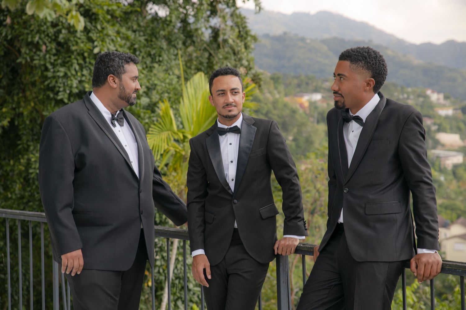 Three men in black tuxedos conversing outdoors with lush green hillside backdrop.