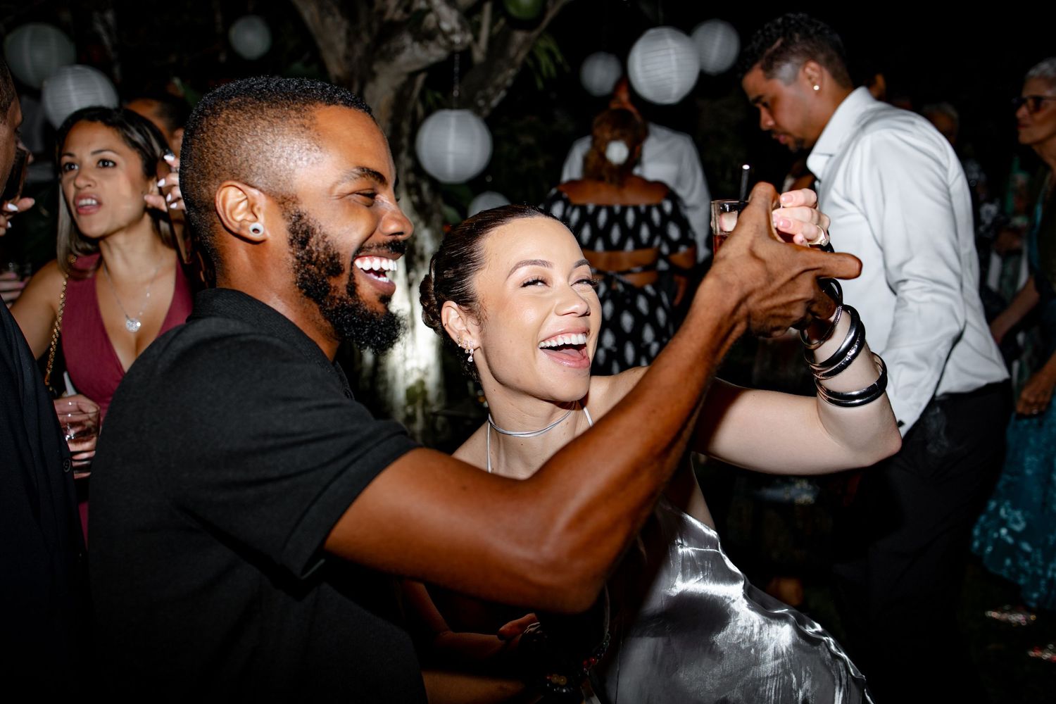 Couple laughs and dances joyfully together at a lively outdoor wedding reception under white lanterns.