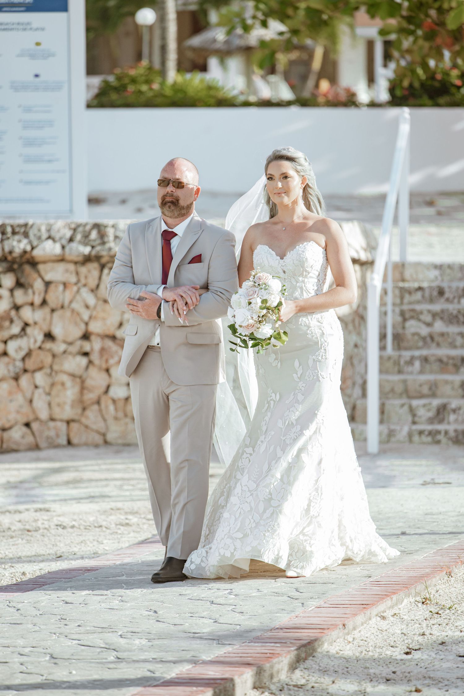 Bride in white lace gown holding bouquet walks down aisle with bald man in tan suit at outdoor wedding.