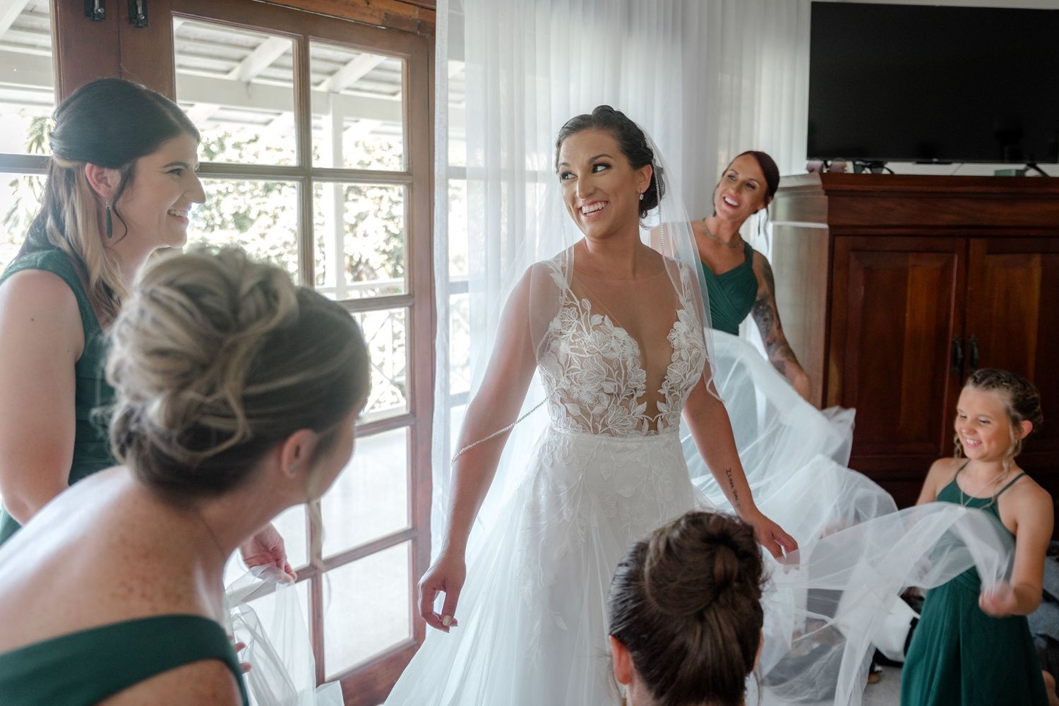 Smiling bride in lace wedding gown surrounded by bridesmaids in green dresses before ceremony.