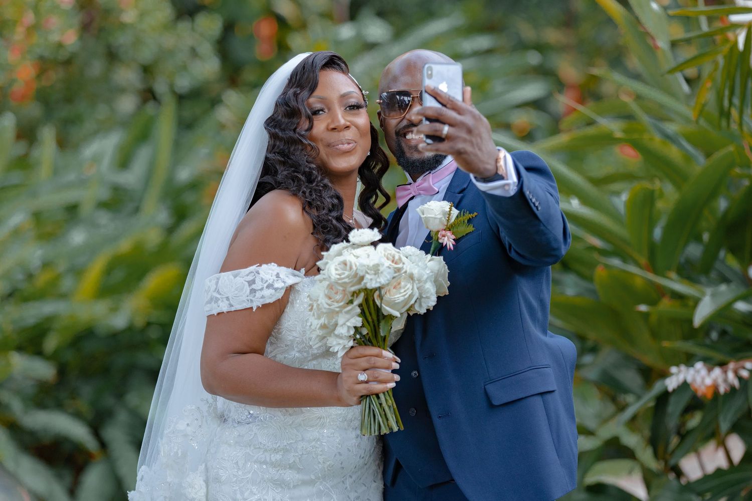 Bride and groom taking a selfie outdoors, bride holding white bouquet in lace gown with veil, groom in navy suit.