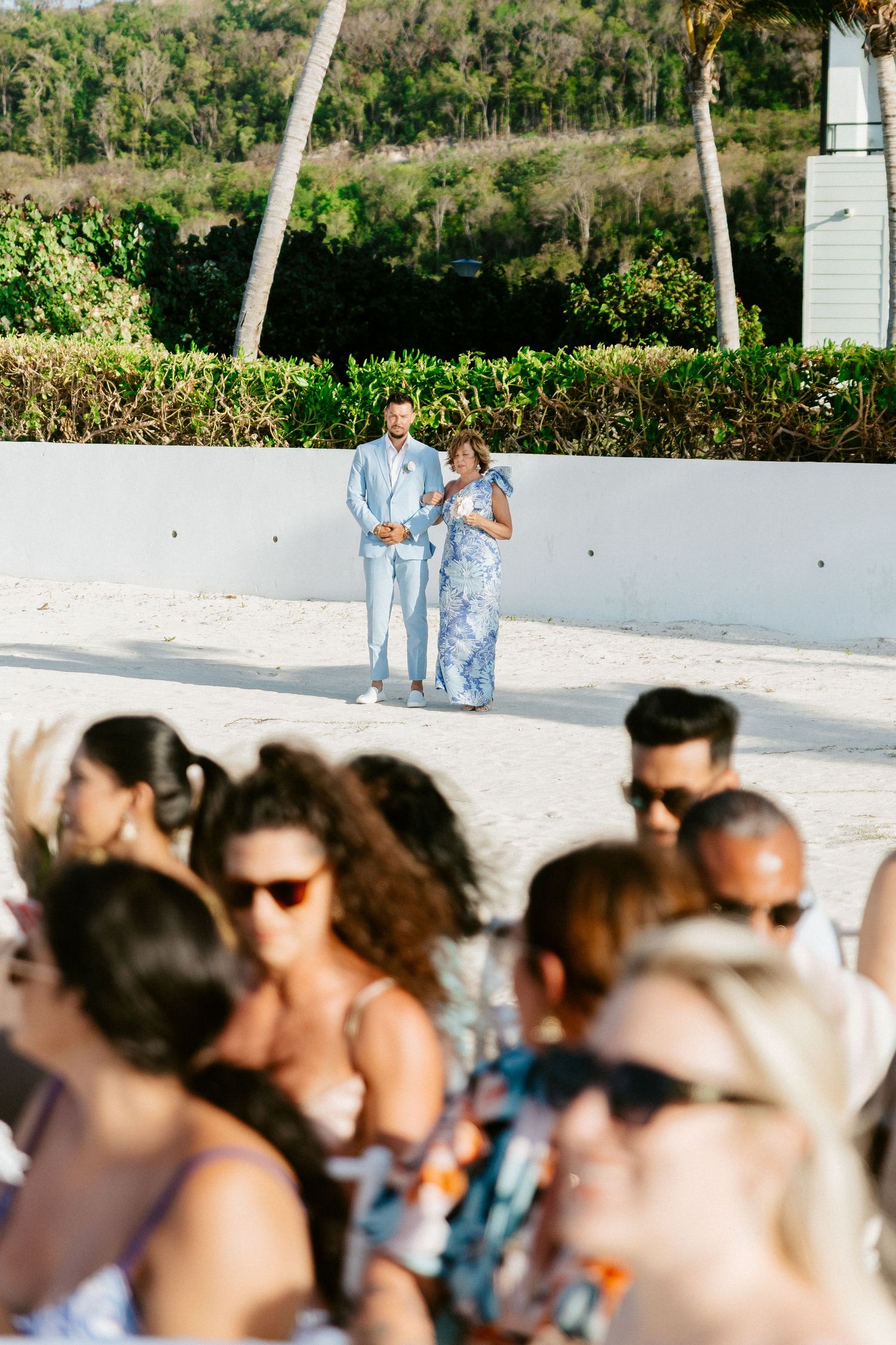 Couple in blue outfits standing before seated wedding guests on a sunny beach ceremony.