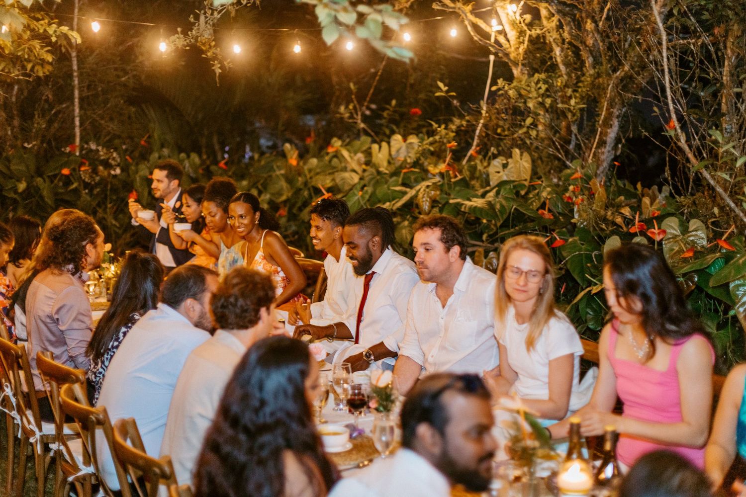 Guests dining at elegant outdoor wedding reception under string lights surrounded by tropical greenery.
