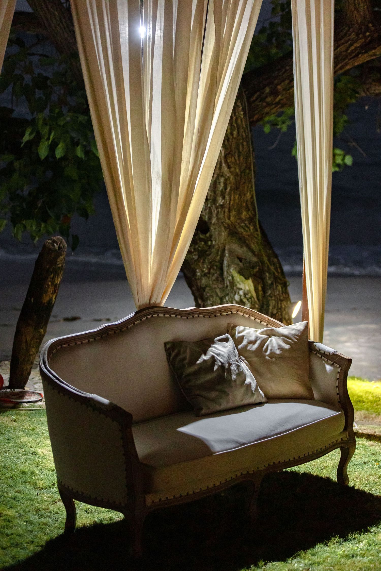Vintage upholstered loveseat with silk pillows under a draped canopy with cream curtains at a nighttime outdoor wedding.