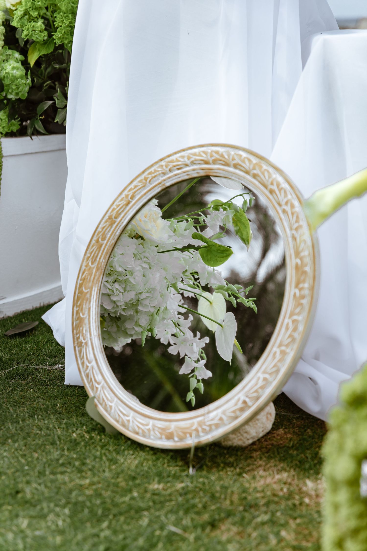 Elegant oval mirror with ornate gold frame reflecting white floral arrangements on green grass at a wedding.