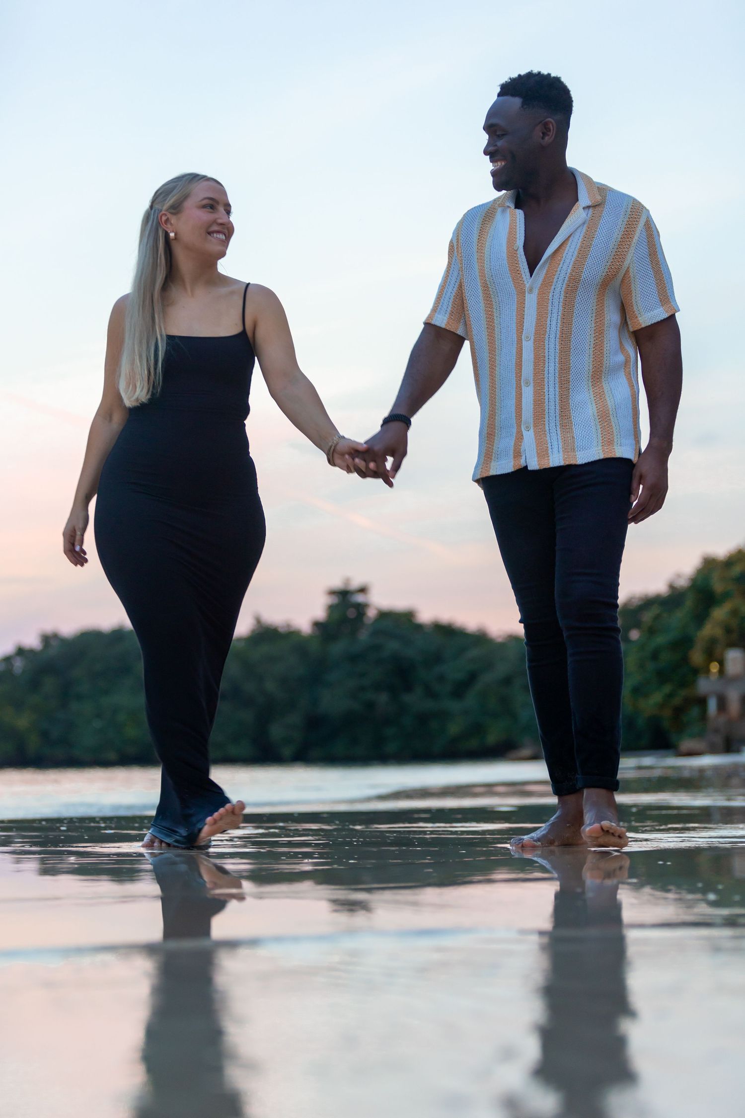 Happy interracial couple holding hands and smiling while walking barefoot on beach at sunset.