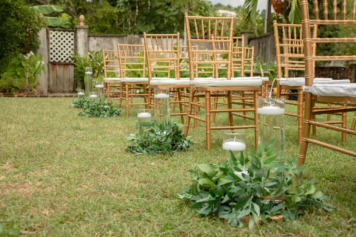 Gold chiavari chairs line garden wedding aisle with glass lanterns and greenery decorations.