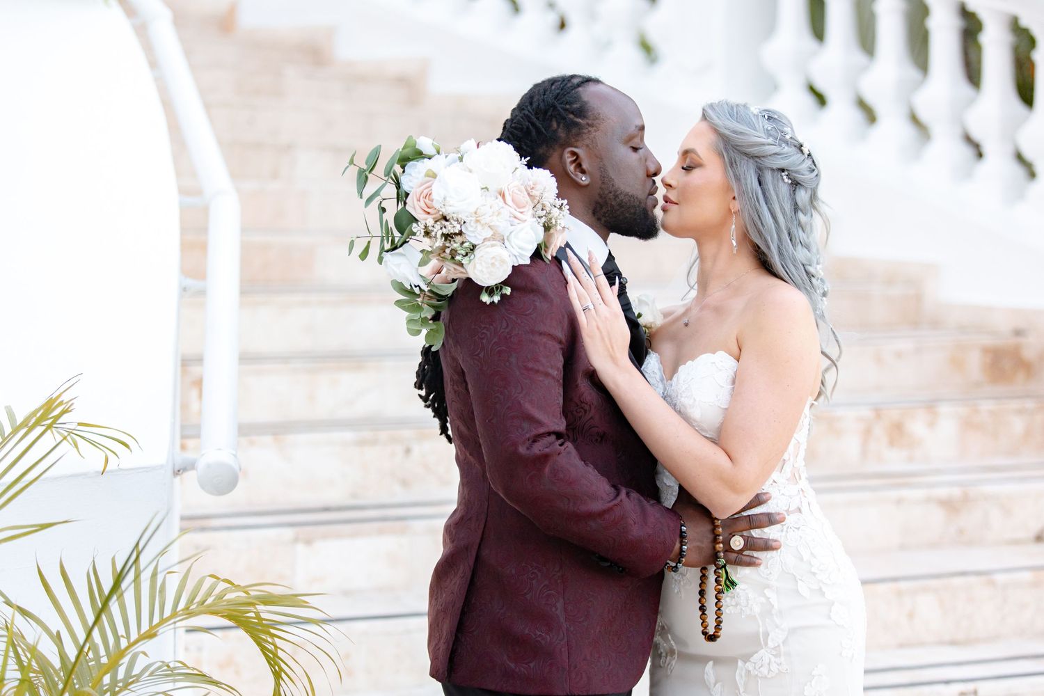 Interracial newlyweds share a kiss while bride holds large blush floral bouquet outdoors.