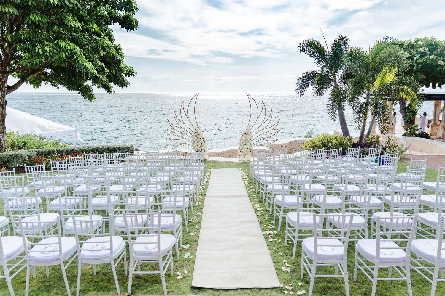 Elegant outdoor oceanfront wedding ceremony setup with white chiavari chairs and golden angel wing altar decor.