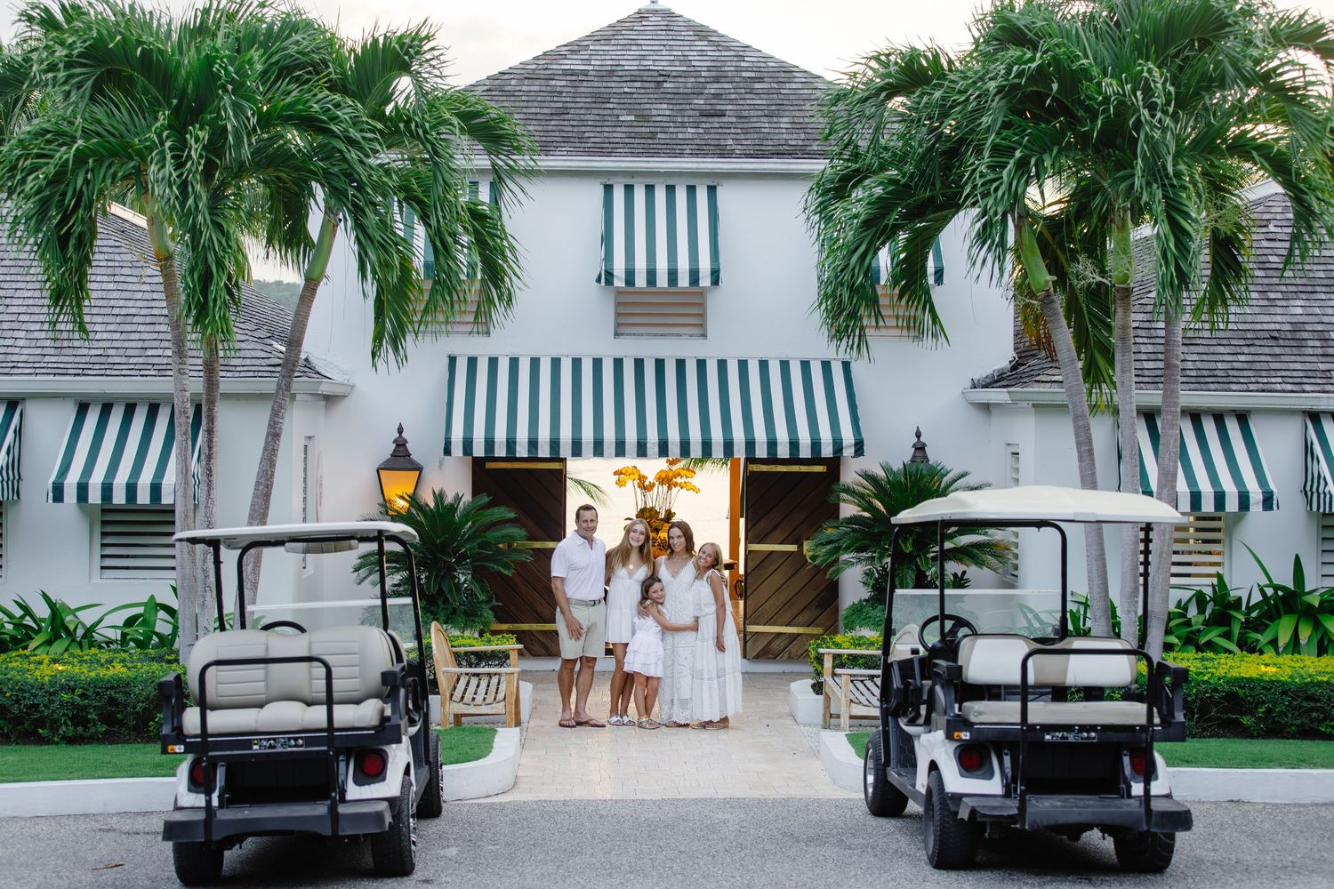 Family in white outfits poses at elegant tropical resort entrance with palm trees and green-striped awnings, golf carts nearby.