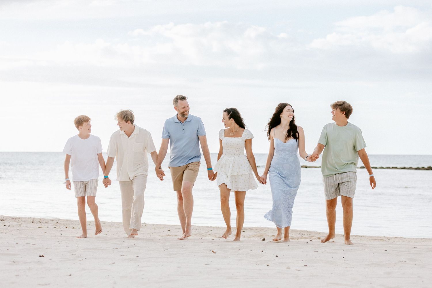 Family of six holding hands walking on a sandy beach with calm ocean waves in the background.