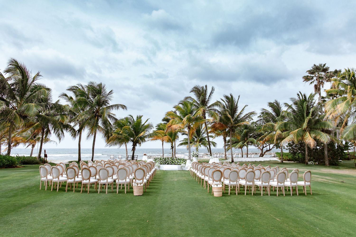 Elegant outdoor wedding ceremony setup on lush green lawn with rows of chairs, palm trees, and ocean view.