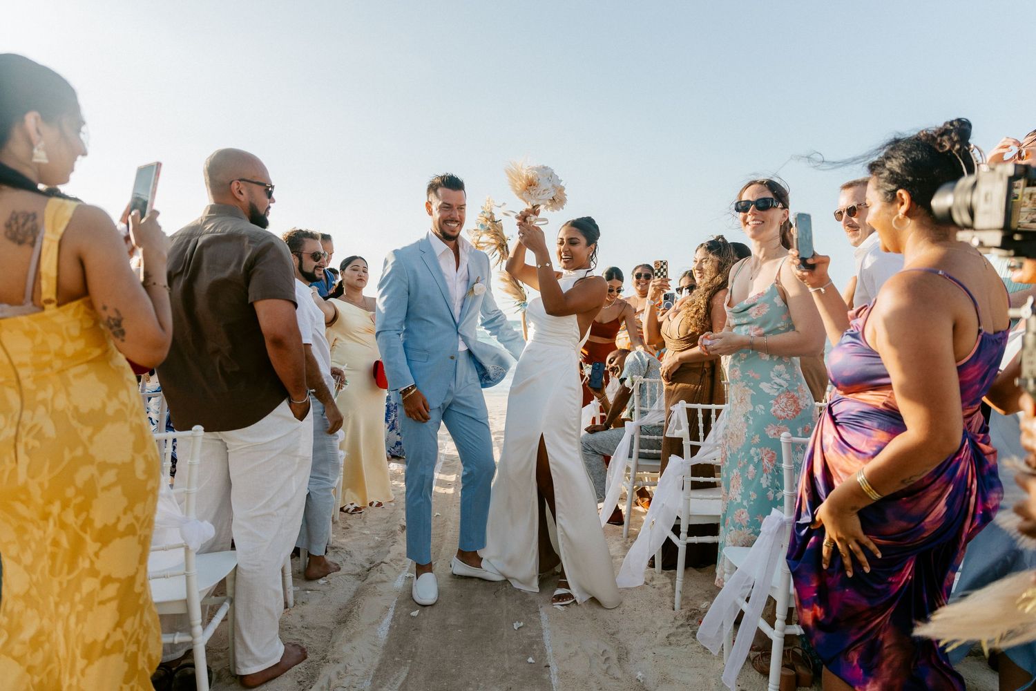 Joyful newlyweds walking down outdoor beach wedding aisle surrounded by cheering guests.