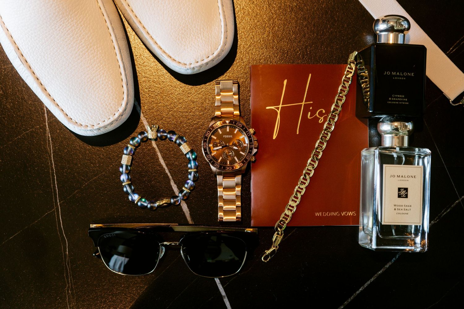 Flat lay of groom accessories including white loafers, gold watch, bracelet, sunglasses, cologne, and a gold chain on dark surface.