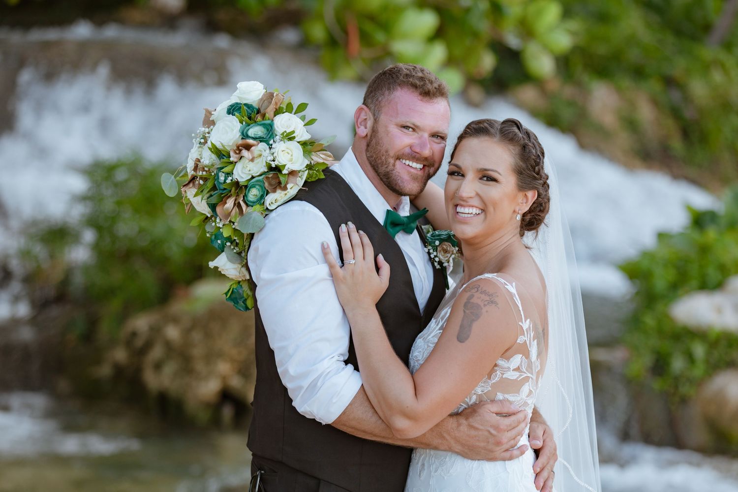 Smiling newlyweds posing together in front of a waterfall, bride holding a lush green and white bouquet.