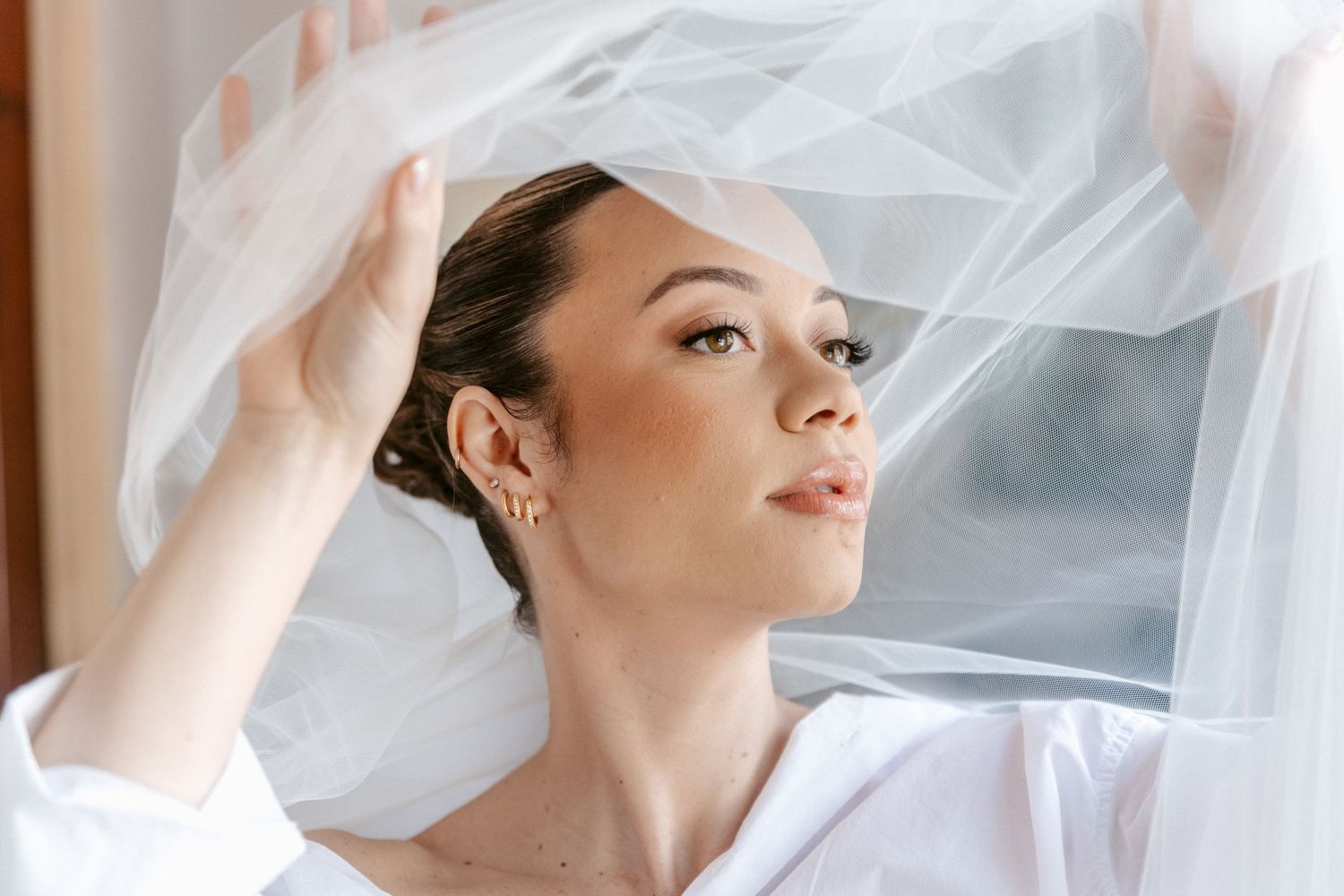 Bride lifting sheer wedding veil with eyes open in soft natural window light.