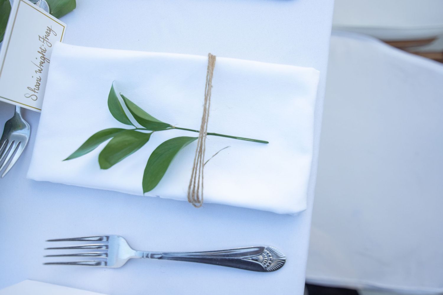 Elegant white wedding place setting with green leaf sprig tied with twine beside silver fork.