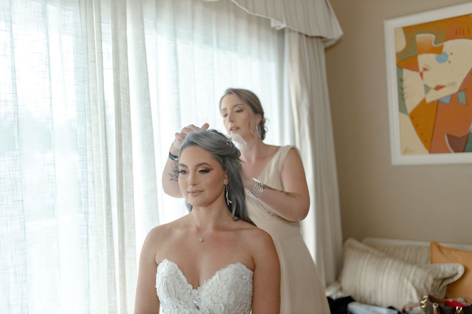 Bridesmaid adjusting silver-haired bride's veil near a bright window before the wedding.