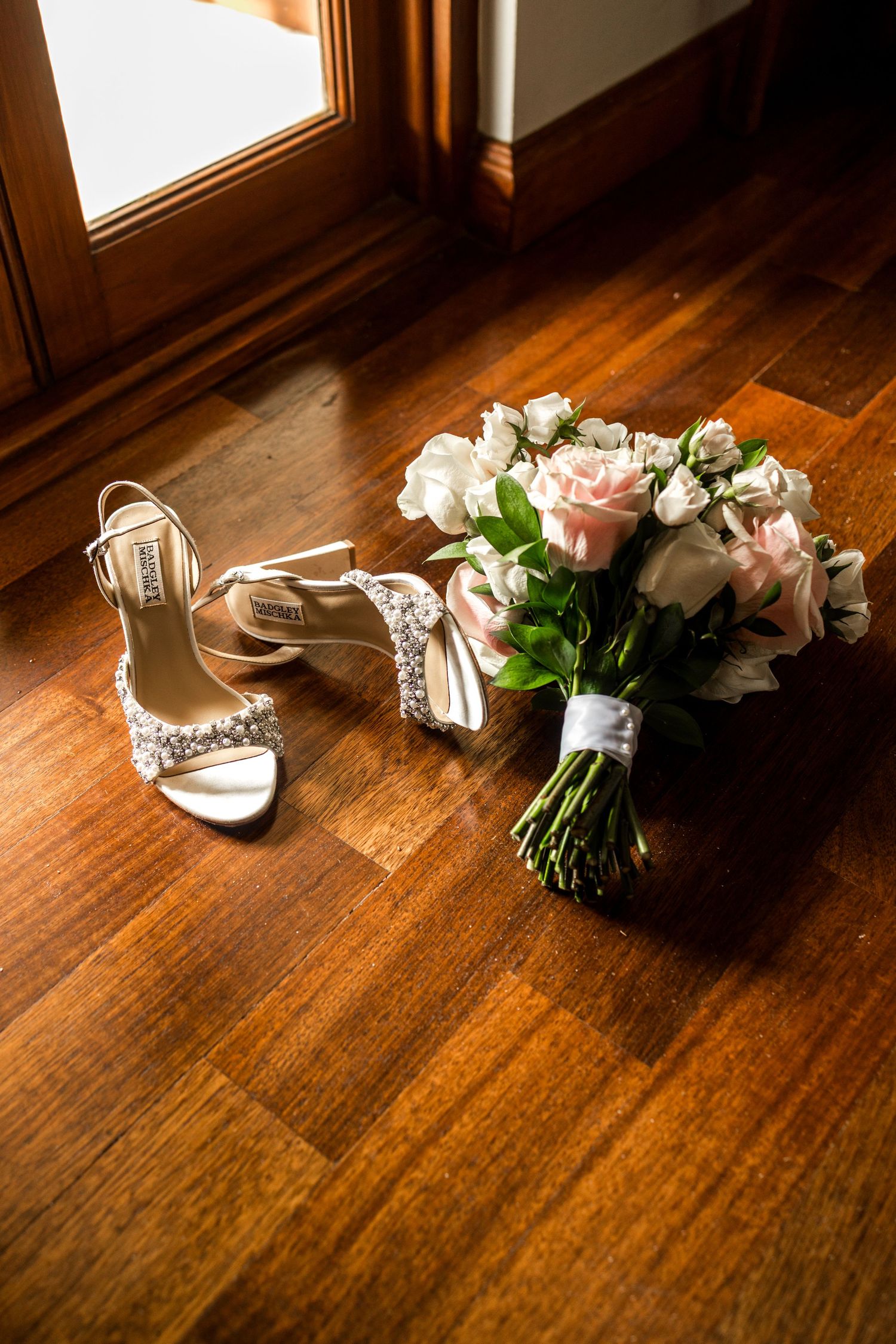 Elegant bridal heels with crystal embellishments beside a pink and white rose wedding bouquet on hardwood floor.