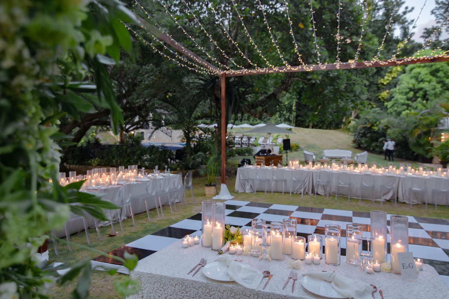 Outdoor wedding reception with long tables, candles, string lights, and a checkered dance floor at dusk.