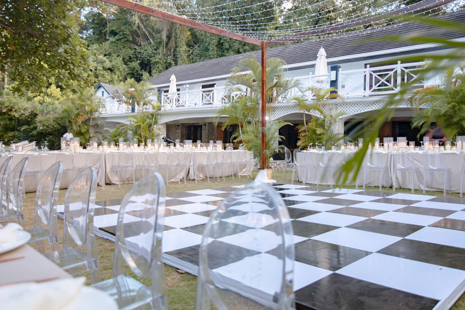 Outdoor wedding venue with black and white checkered dance floor, ghost chairs, and white reception tables beside an elegant estate.