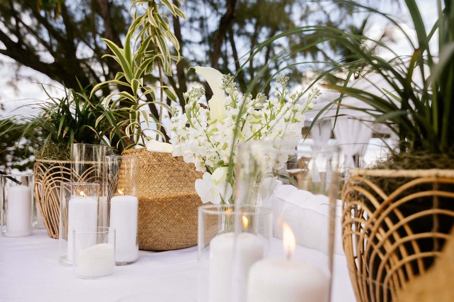 Elegant outdoor wedding table with white candles, tropical greenery, white orchids, and woven rattan accents on a white tablecloth.