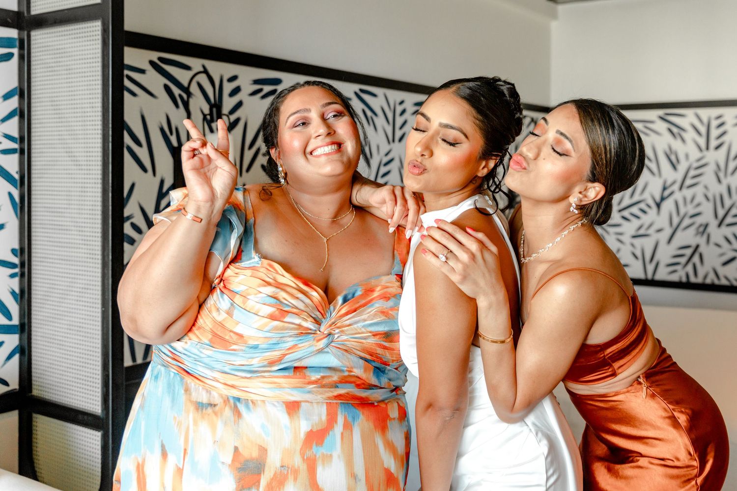Three women in colorful formal dresses pose together, smiling and making fun gestures at a wedding celebration.