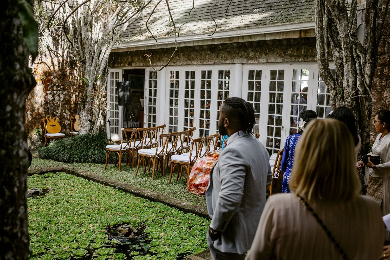 Wedding guests viewing outdoor reception setup with white French doors and wooden chairs on garden lawn.