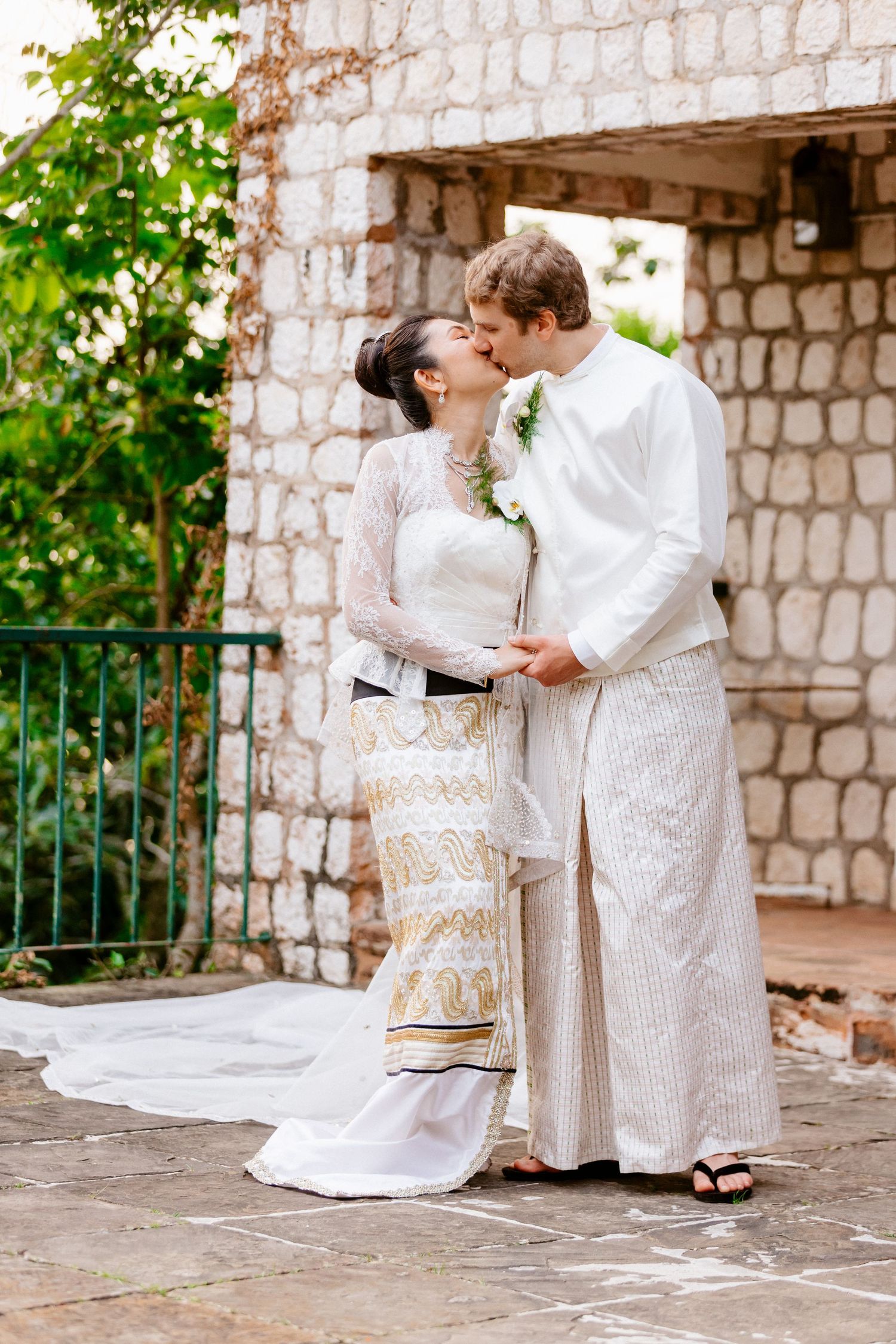 Newlywed couple sharing a kiss in traditional white wedding attire at rustic stone outdoor venue.
