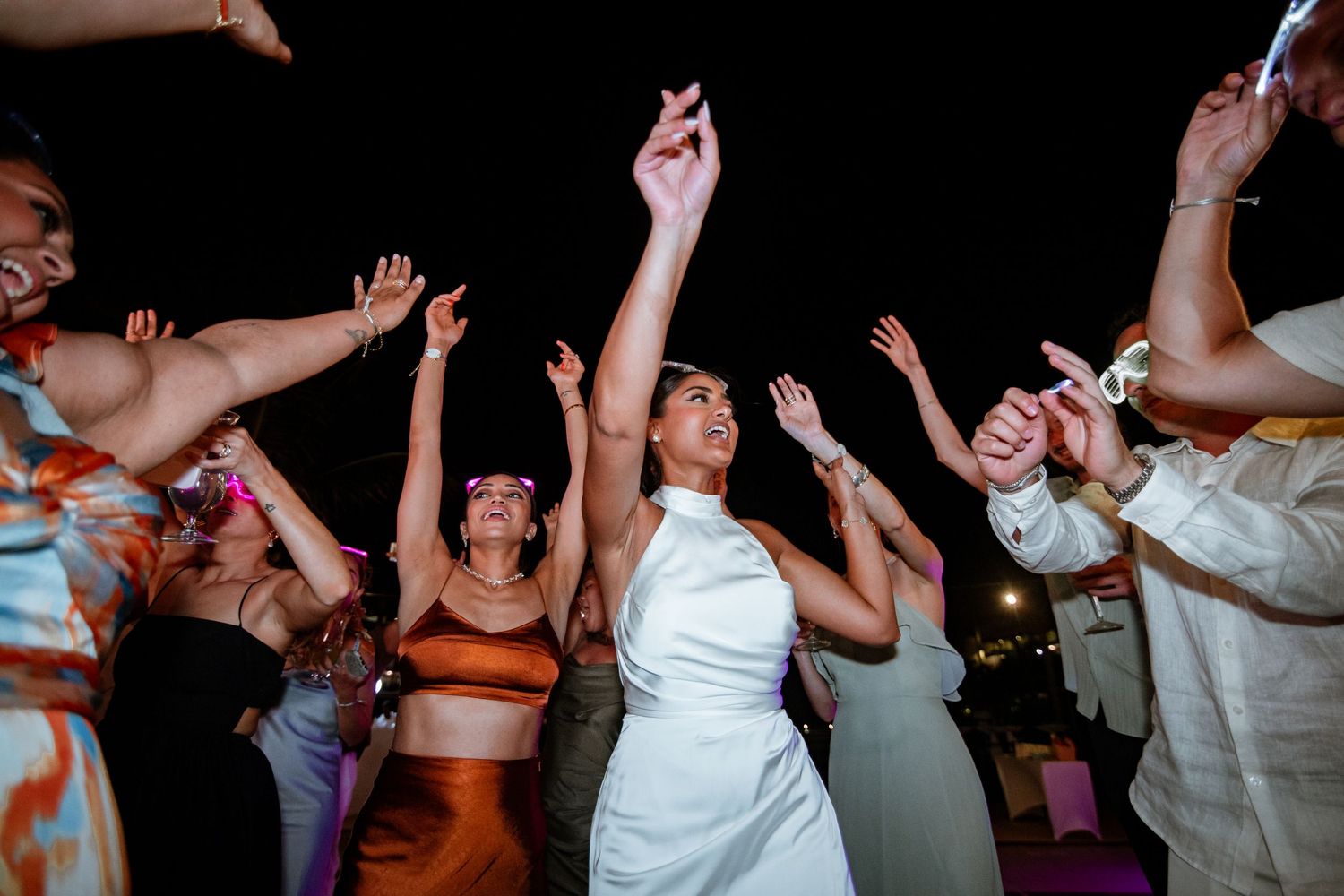 Bride in white halter dress dancing with guests under purple lights at lively outdoor wedding reception.