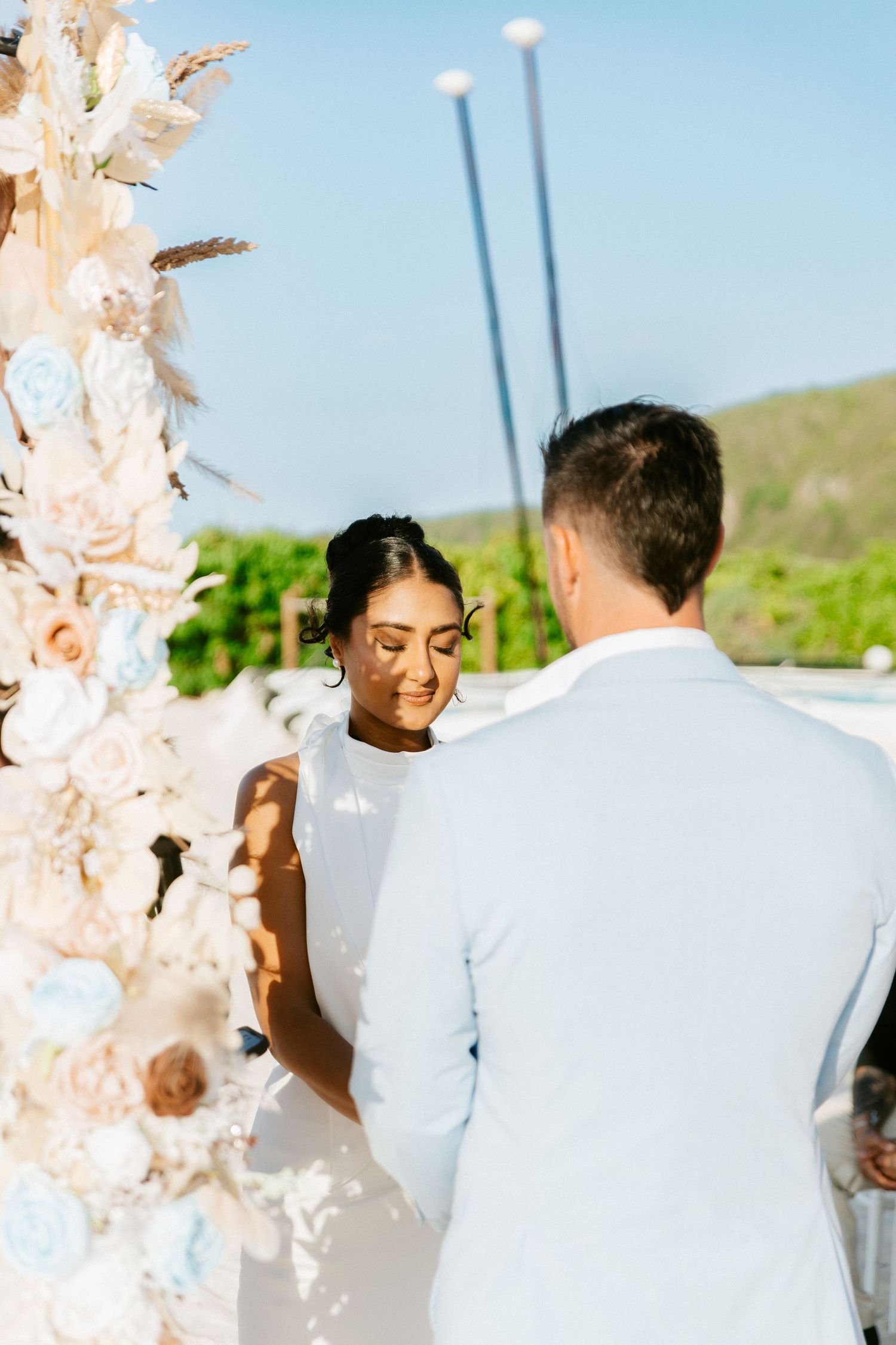 Bride and groom exchange vows at a floral arch adorned with white and blush flowers during an outdoor ceremony.