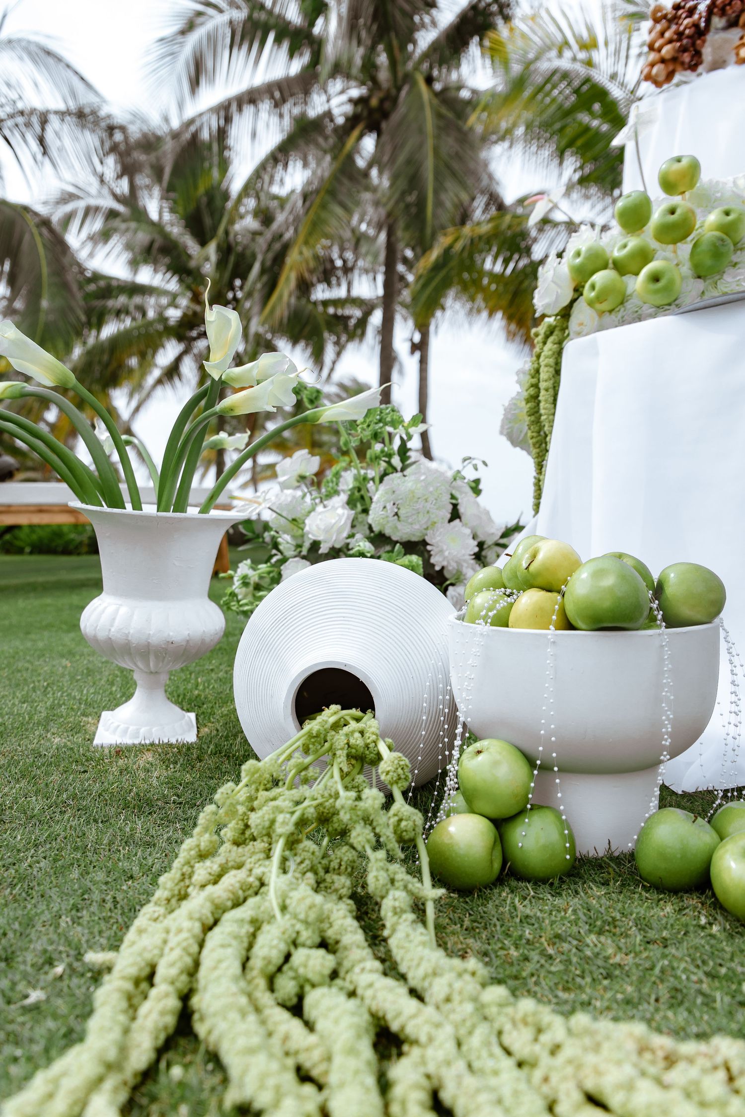 White vases with green apples, limes, and floral arrangements on grass with tropical palm trees backdrop.