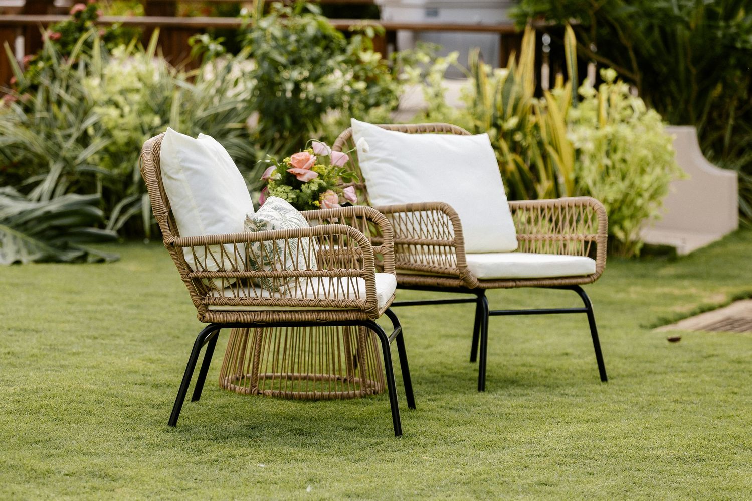 Two rattan outdoor chairs with white cushions on green grass, surrounded by lush garden plants and a floral centerpiece.