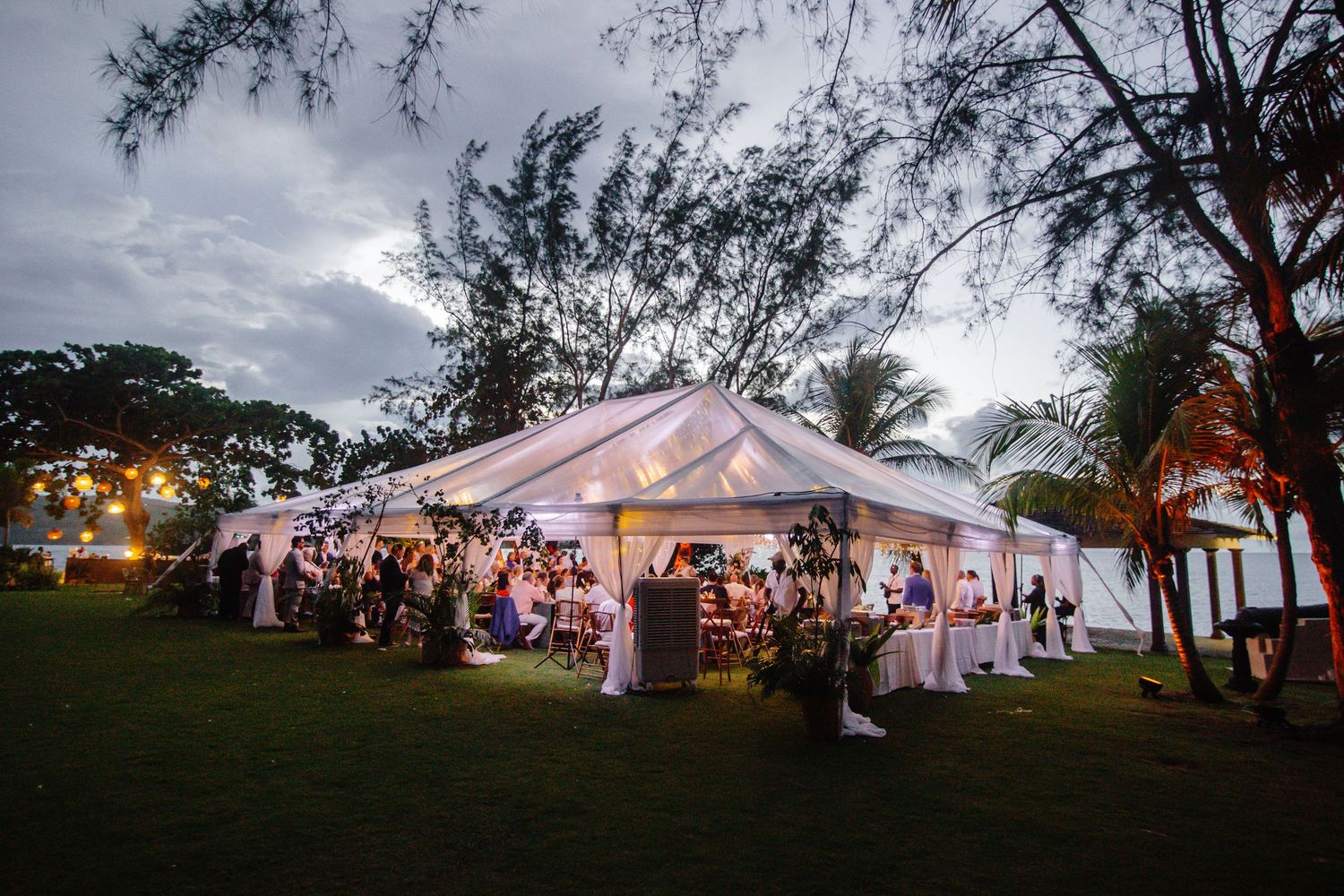 Elegant outdoor wedding reception under a white tent with warm lighting at dusk, surrounded by tropical trees and lush green lawn.